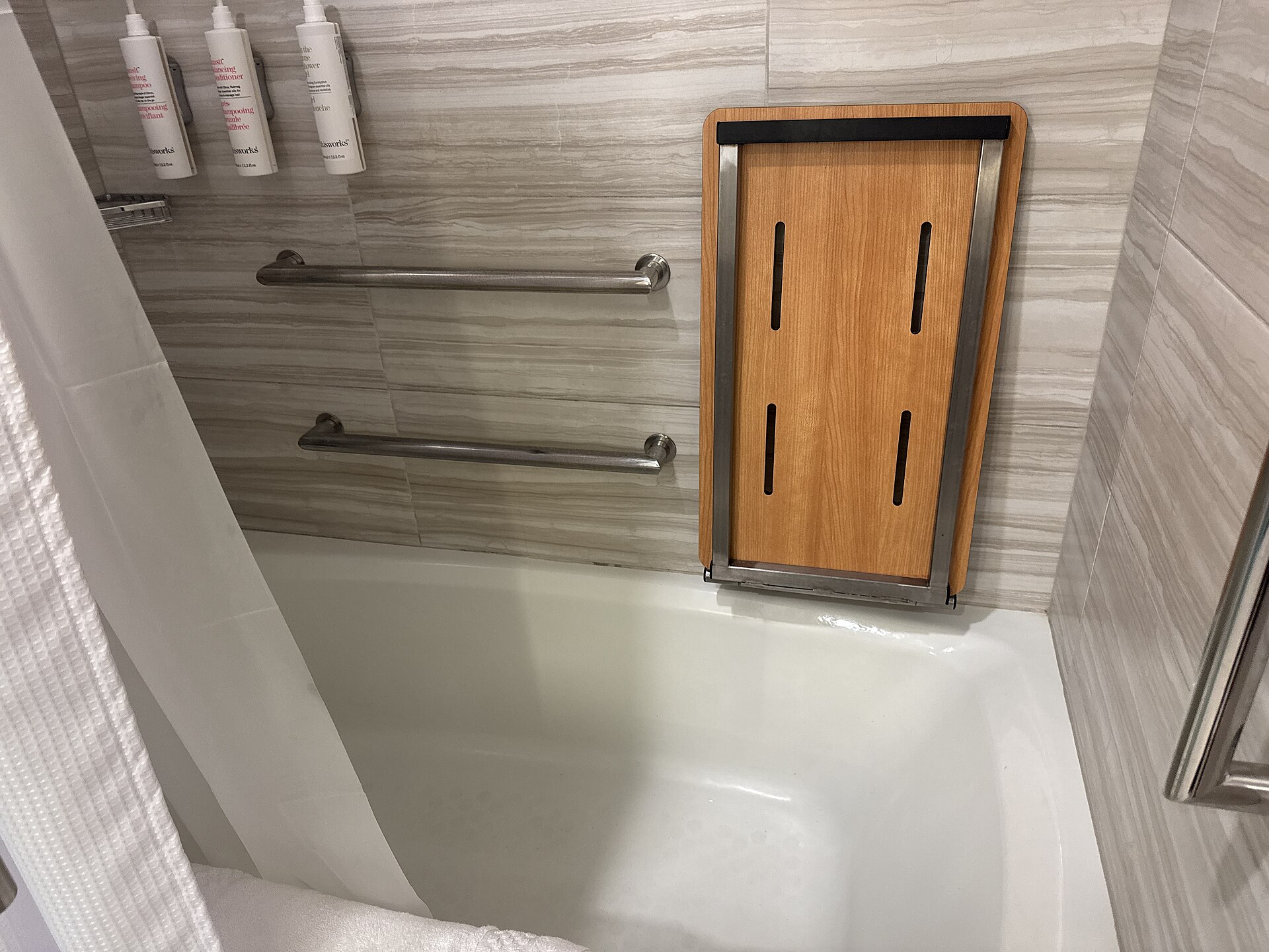 Bathtub with two horizontal grab bars and a fold-down wooden shower seat on a tiled wall