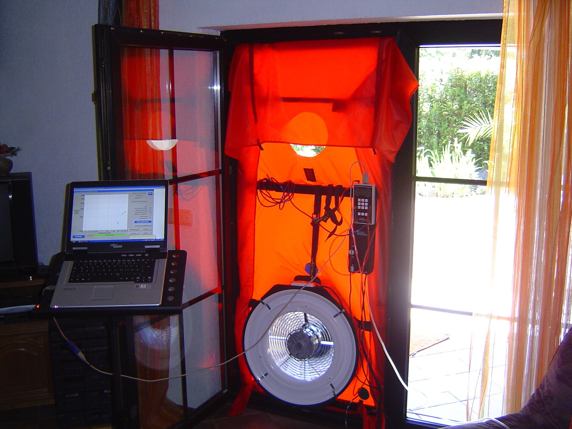 Blower door test rig with a fan in a red frame fitted into a patio doorway, connected to a laptop