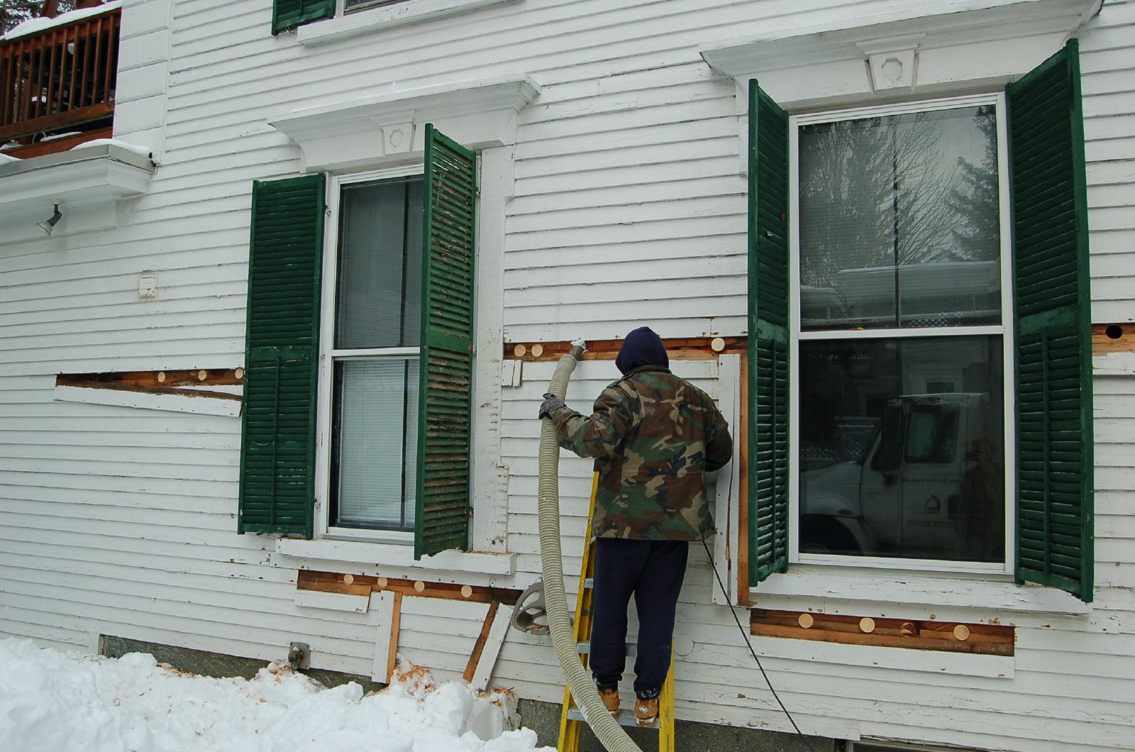 Installer standing in snow blowing cellulose insulation into the exterior wall of a white clapboard house