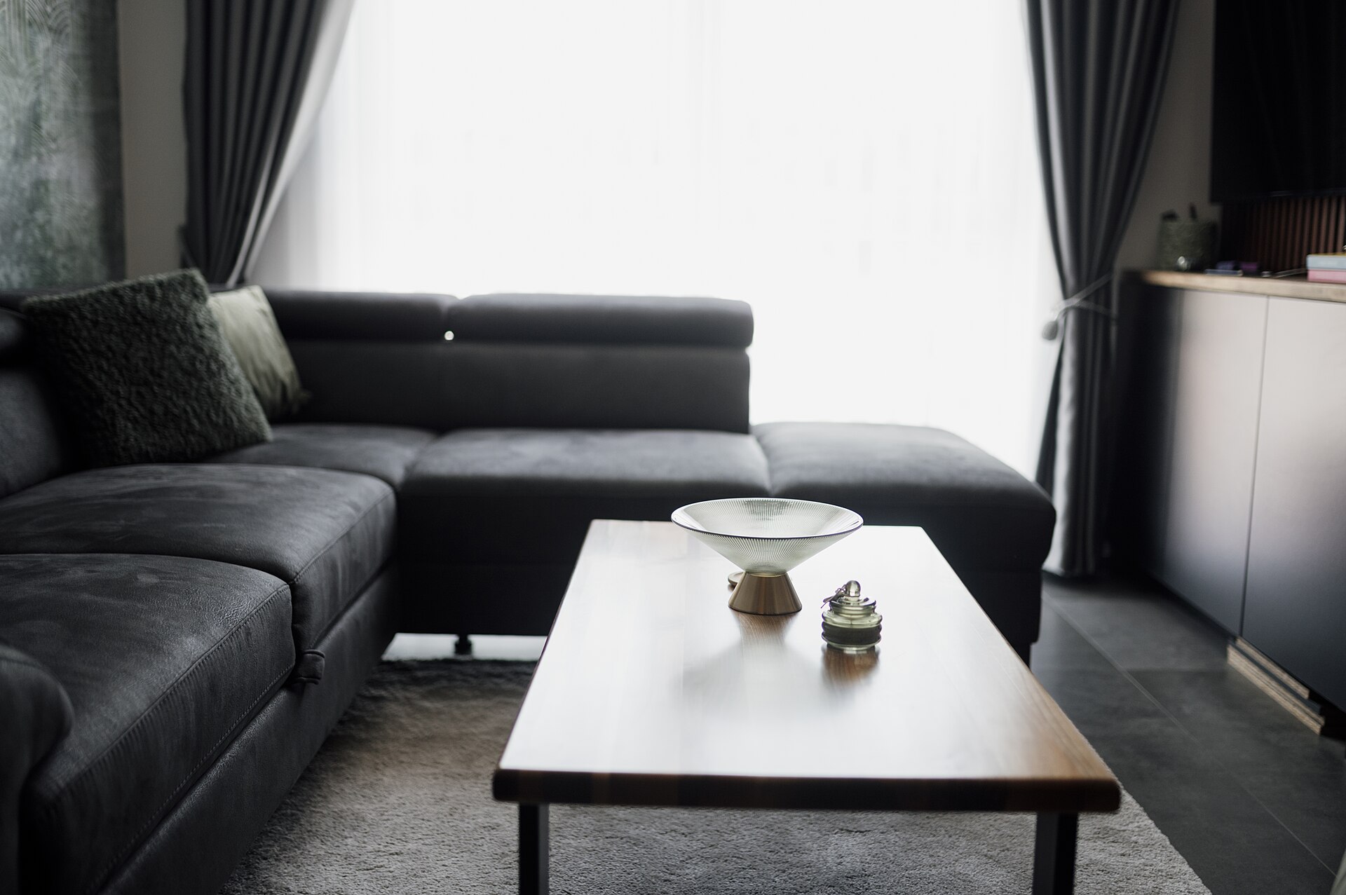 Simple living room with a grey sectional sofa and low coffee table lit by a bright, sheer-curtained window