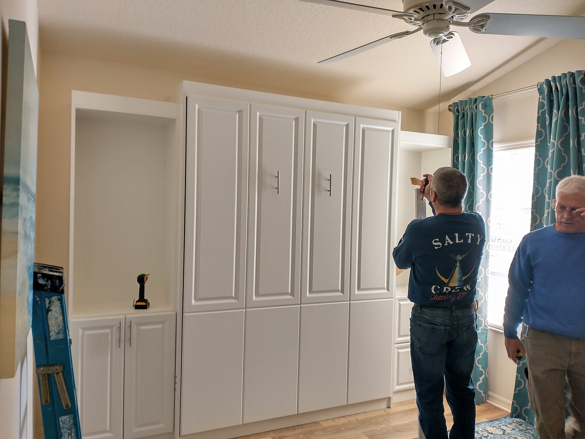 Installers fitting a white built-in wall bed cabinet with flanking shelving units in a bedroom