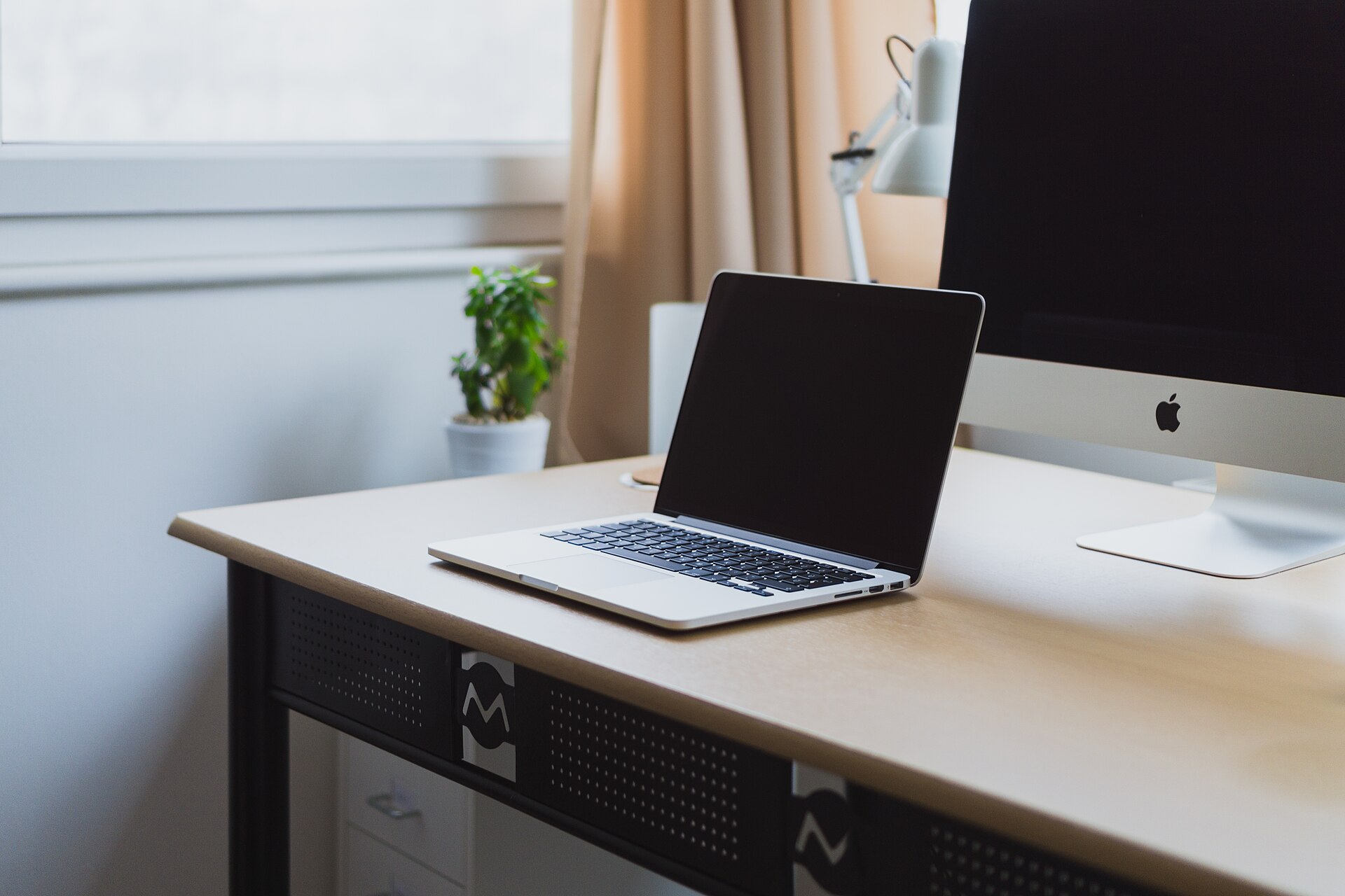 Laptop and desktop monitor on a tidy desk beside a window with a small potted plant