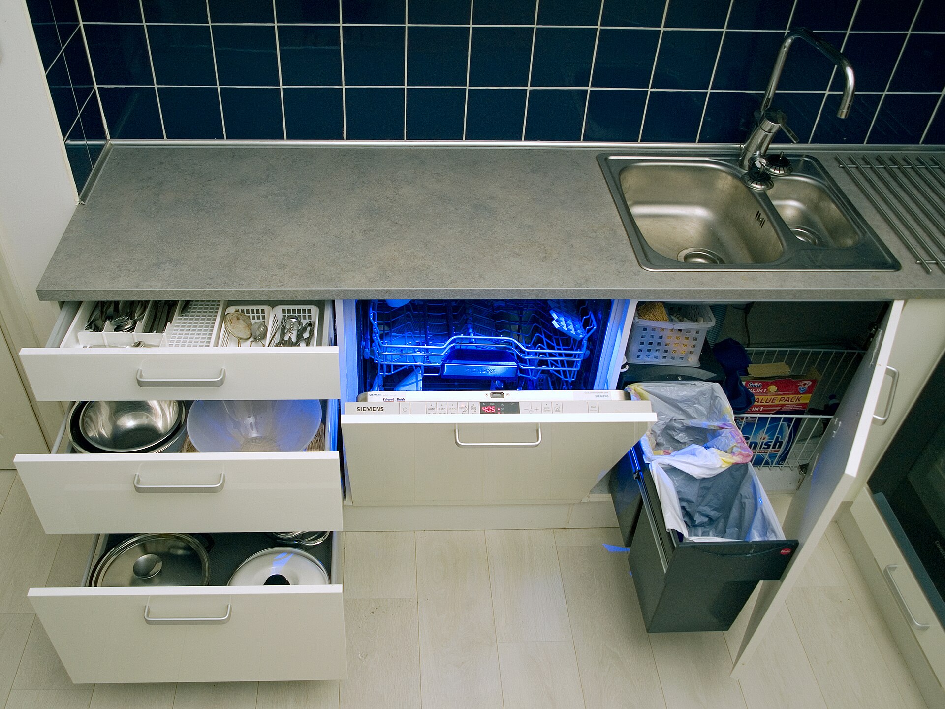 White kitchen lower cabinets open showing pull-out drawers for pots, a dishwasher and a pull-out bin