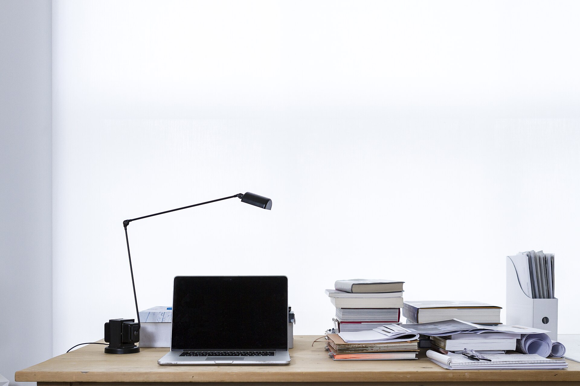 Minimal white desk with a laptop, desk lamp, stacked books and a pencil cup against a white wall