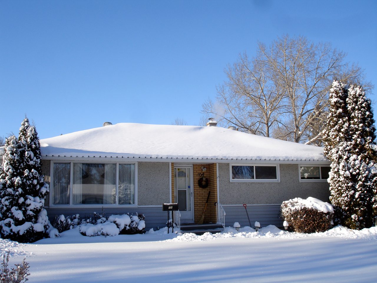 Single-storey bungalow with a snow-covered roof, evergreen shrubs and a snowy front yard on a sunny winter day