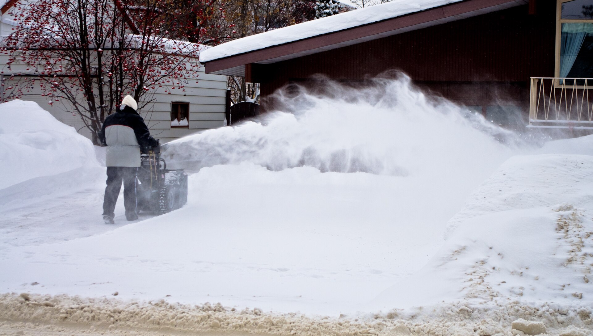 Person using a two-stage snowblower to throw deep snow off a driveway beside a garage