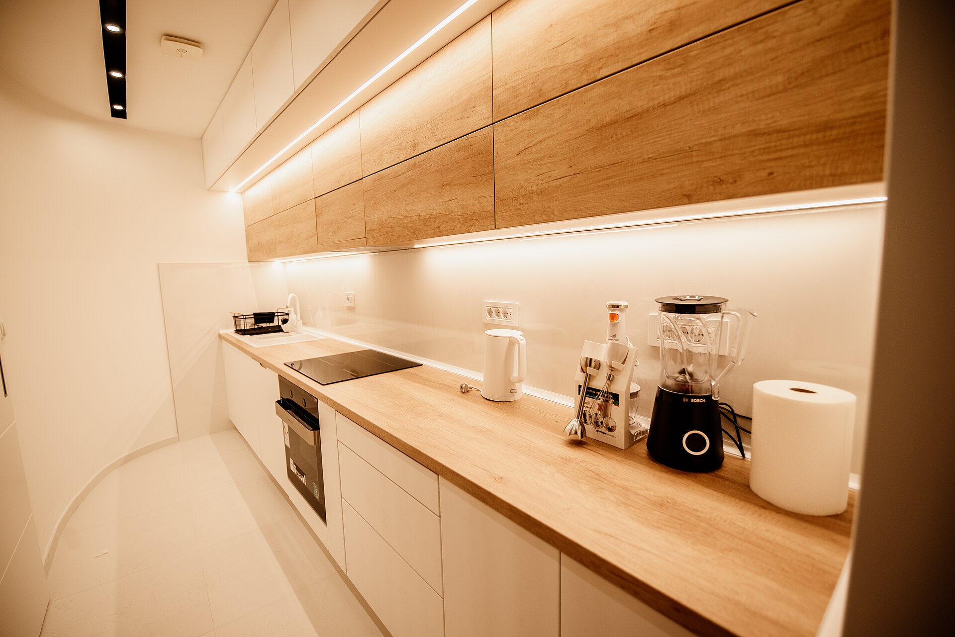 Narrow modern kitchen with white lower cabinets, wood counters and LED strip lights under the upper cabinets