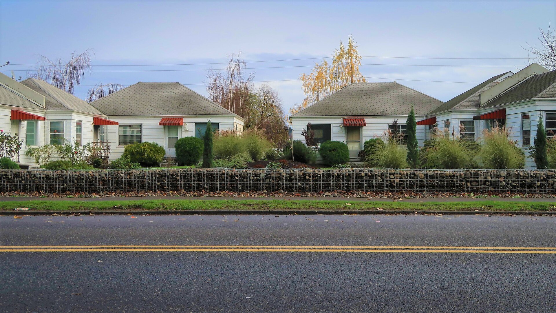 Group of small single-storey cottages with gabled roofs sharing one landscaped lot behind a low fence