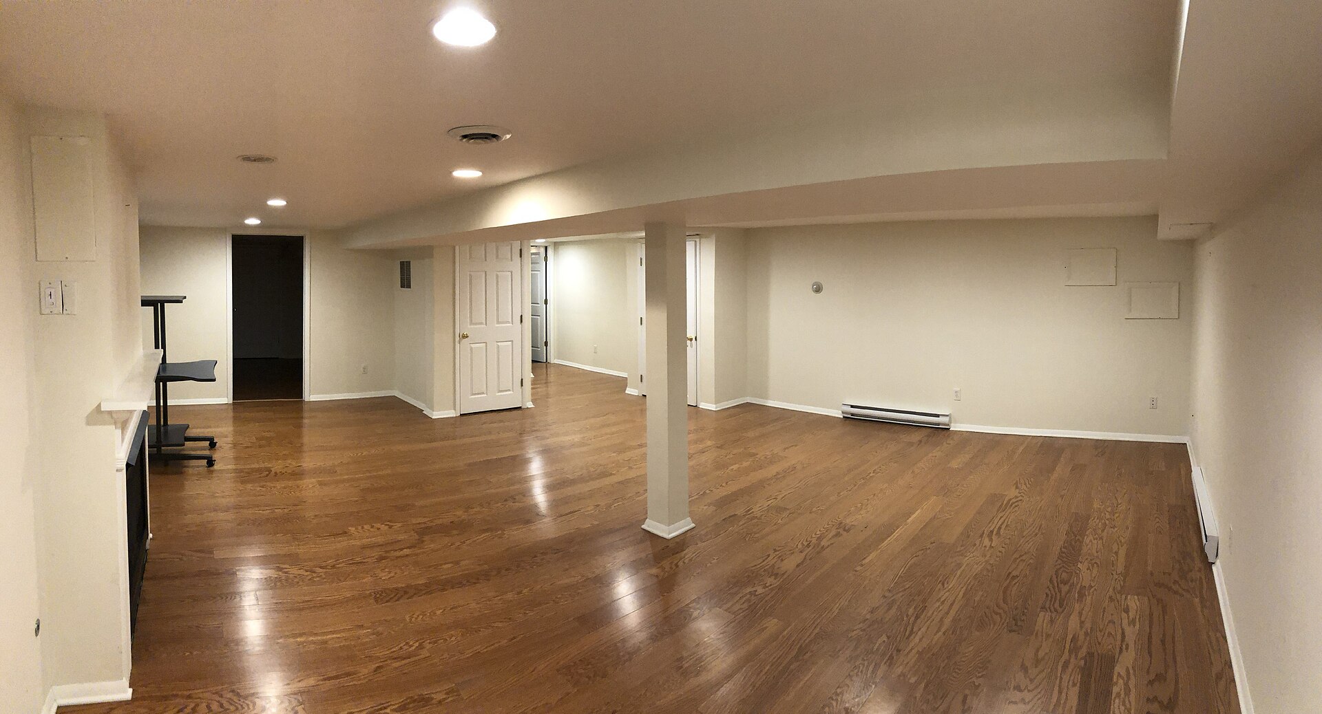Wide view of a finished basement with hardwood floors, pot lights and white support posts
