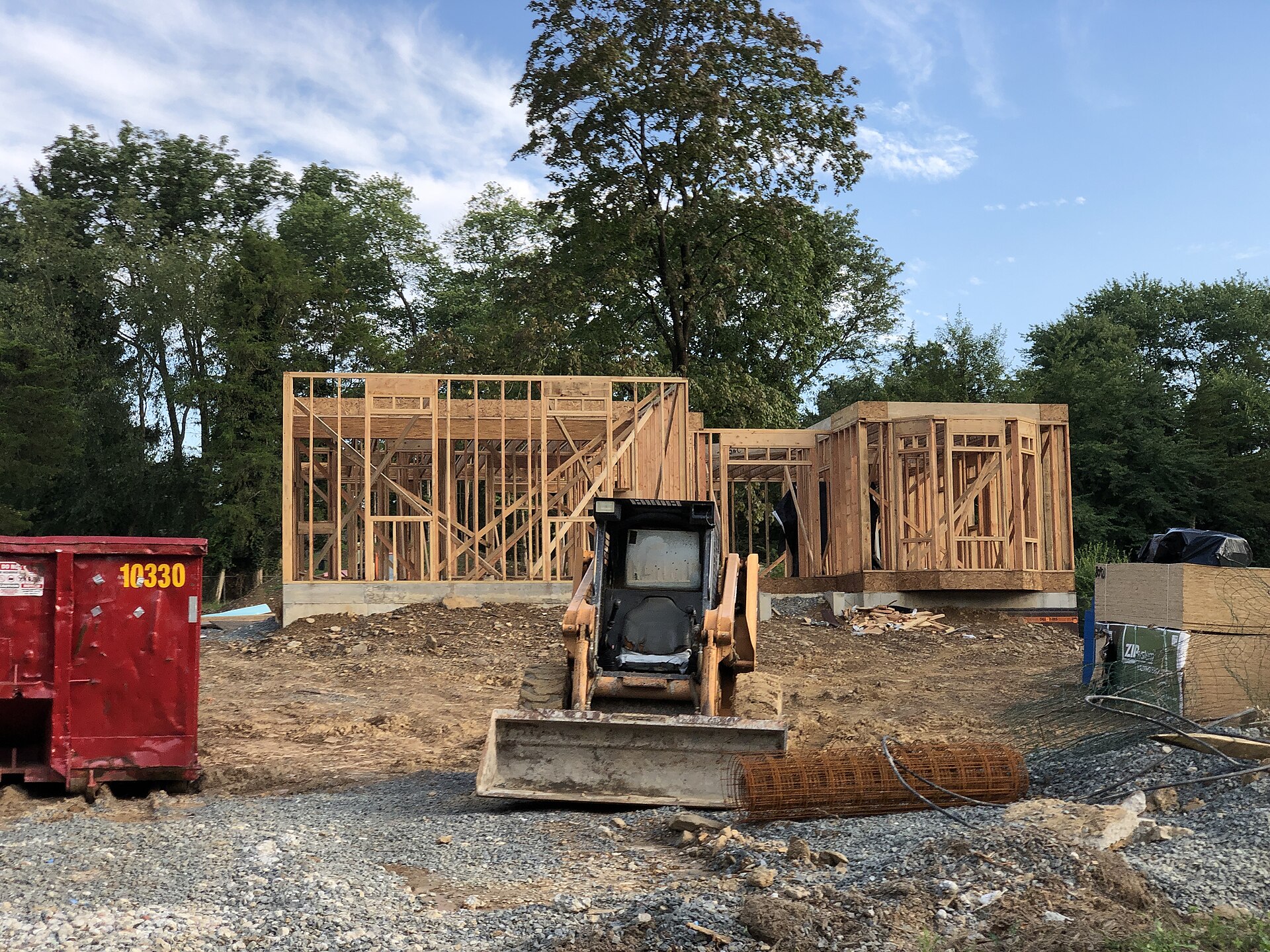Wooden frame of a new house rising on a dirt lot with a skid steer loader and dumpster in front