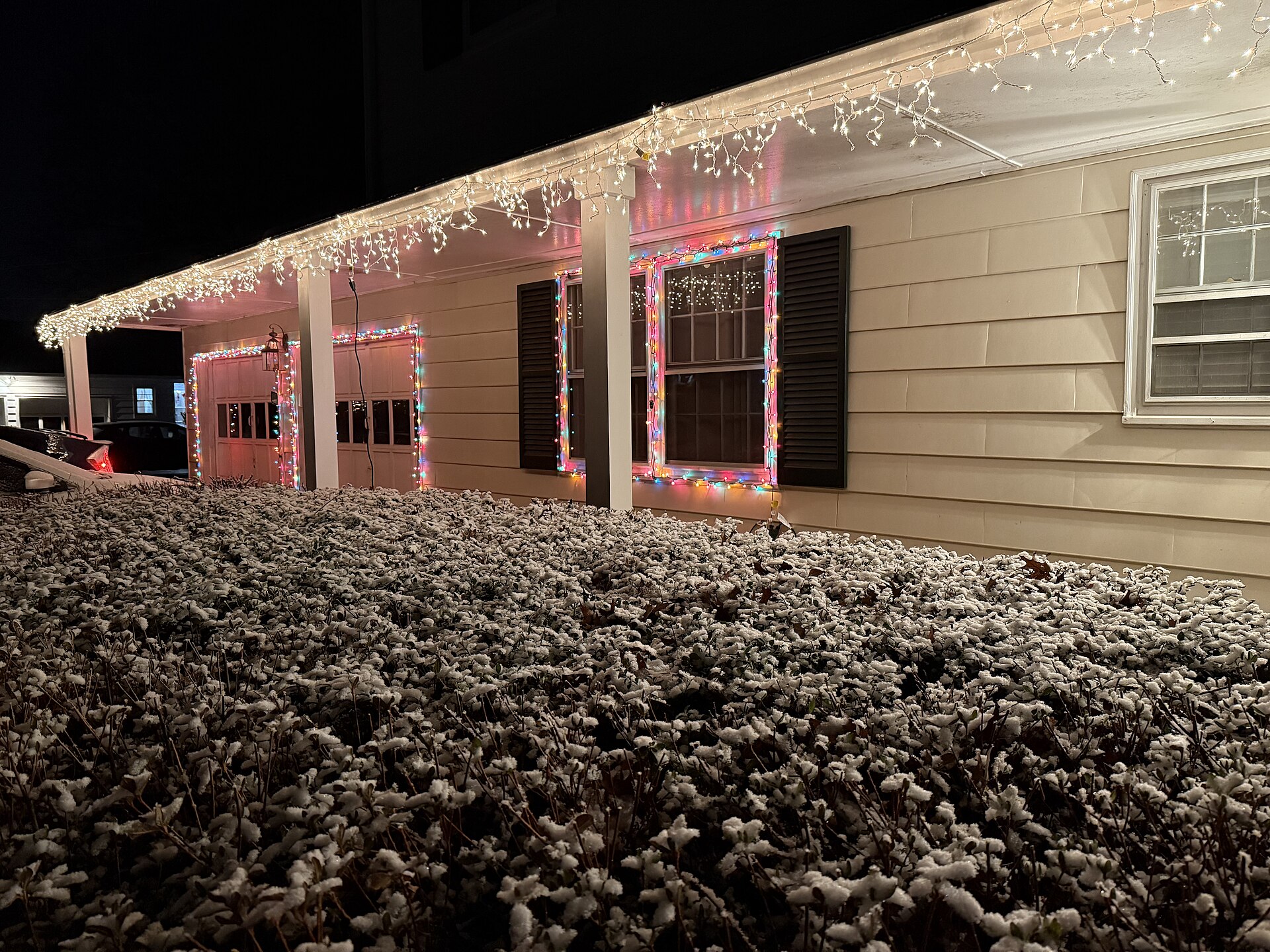 Icicle-style string lights along the eaves and coloured lights around windows of a house above snow-dusted shrubs
