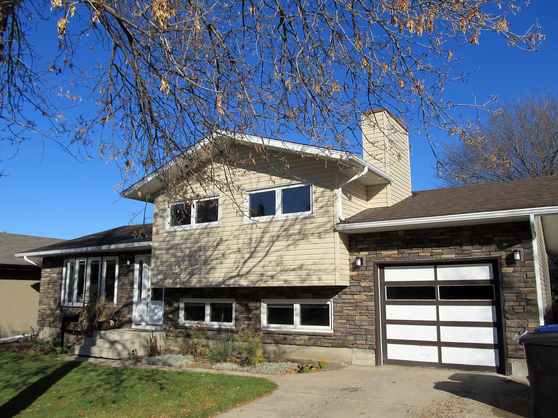 Split-level house in Saskatoon refinished with tan vinyl siding, stone veneer and a modern glass garage door